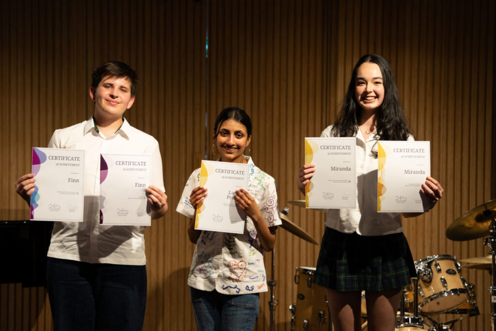 Three students stand on stage, smiling and holding certificates during a Talent Showcase. They wear casual and school clothing. The background features wooden panelling and part of a drum kit. Stage lighting highlights the students, emphasising this moment of student celebration.