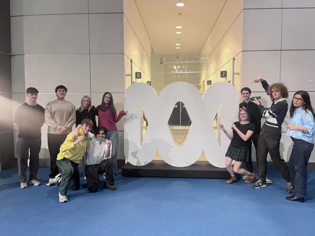 Twelve people pose and smile around a large white abstract sculpture in an indoor space with blue carpet and well-lit walls at the ABC National Forum.