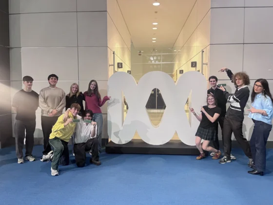Twelve people pose and smile around a large white abstract sculpture in an indoor space with blue carpet and well-lit walls at the ABC National Forum.