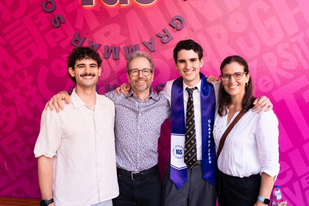 Four people pose smiling in front of a pink wall, one wearing a graduation sash reading Class of 2026. The group includes two men, a graduate in a suit, and a woman wearing glasses.