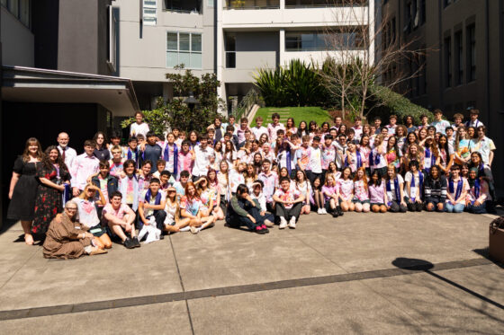 A large group of people, mostly teenagers, pose together outdoors in front of a modern building, many wearing colourful shirts and sashes, smiling at the camera.