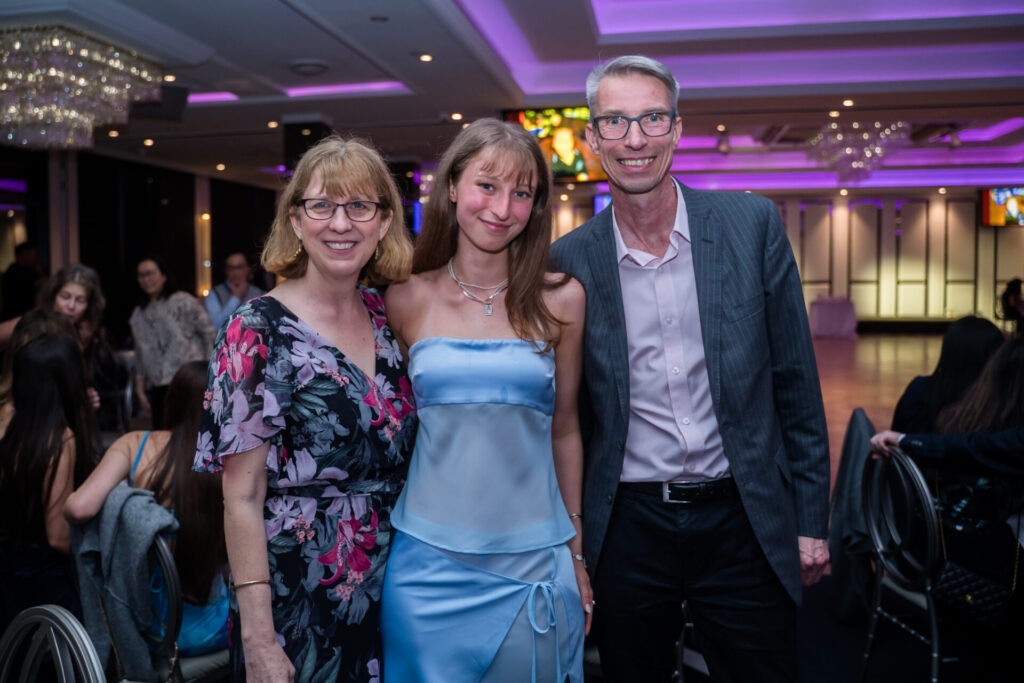 Three people posing and smiling at an indoor graduation event with purple lighting; the woman on the left wears a floral dress, the young woman in the centre—part of the class of 2026—wears a blue satin dress, and the man on the right wears a suit, celebrating this special farewell occasion.