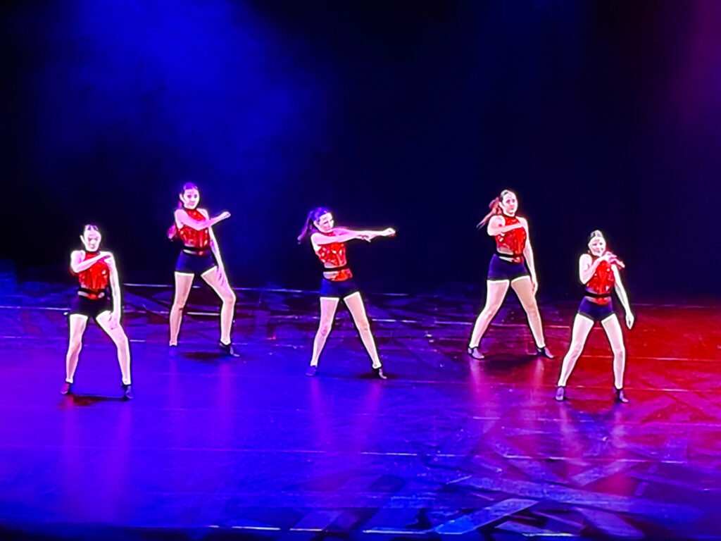 Five dancers in red and black costumes perform onstage under purple and blue lighting, each striking a different energetic pose with arms and legs extended, capturing the vibrant spirit of the forthcoming Independent Dance Festival 2026.