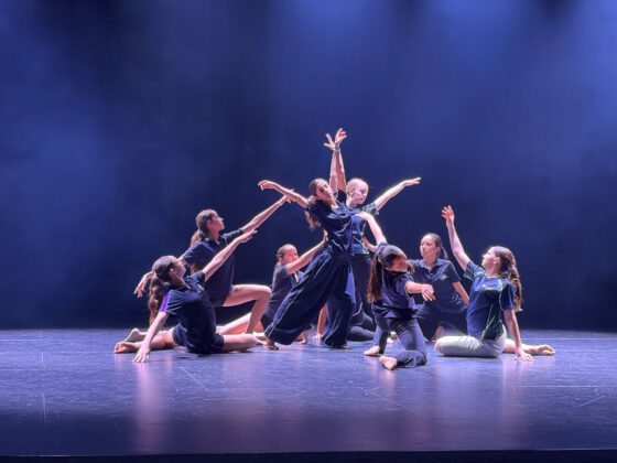 Eight dancers pose dramatically on a stage under blue lights at the Independent Dance Festival, some seated and some reaching upwards with extended arms, creating an expressive, artistic scene.