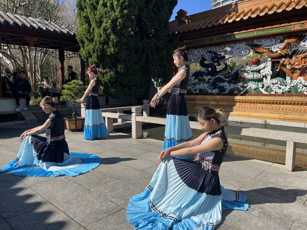 Four women in blue, black, and white dresses perform a dance outdoors in a sunny courtyard with traditional Asian architecture and detailed decorations in the background, highlighting a beautiful cultural connection that celebrates Chinese culture.