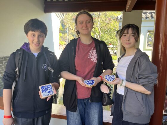 Three young people stand side by side outdoors, smiling and holding up decorative blue and white ceramic tiles, celebrating a cultural connection with Chinese culture. Trees and traditional architecture are visible in the background.