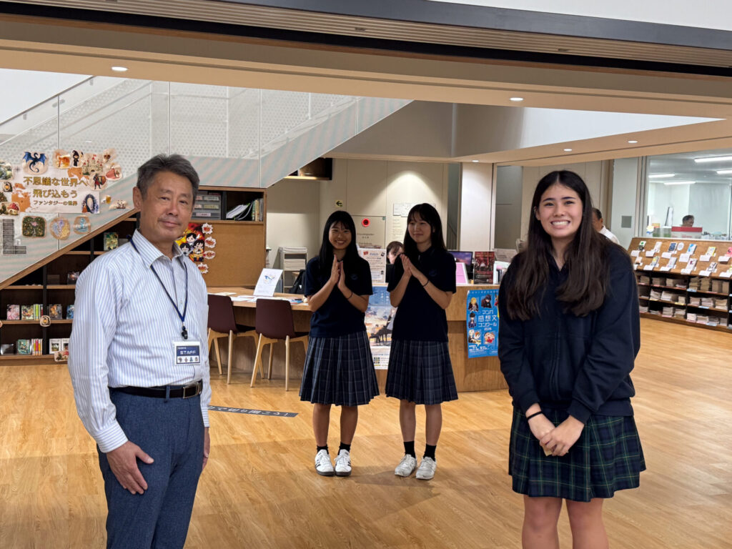A man in a striped dress shirt and three pupils in school uniforms standing together in a library foyer, sharing a sense of friendship and culture.