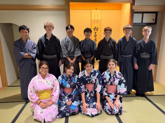 Twelve students in traditional Japanese yukata share a moment of friendship while posing in a tatami mat room, with seven standing in the back row and four kneeling in the front.