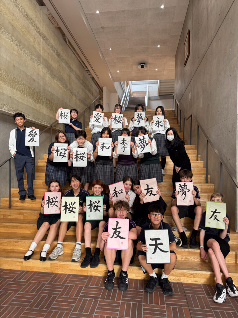 A group of students united by their cultural travel sit and stand on indoor stairs, each holding a piece of paper displaying a Japanese kanji character.