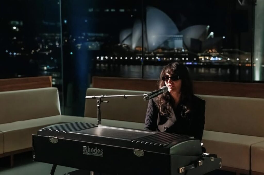 A musician wearing dark glasses plays a Rhodes electric piano for a WITWAT Tripleplay performance, with a microphone set up and the Sydney Opera House visible through a large window at night.