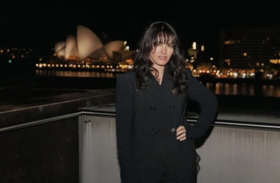 A woman in a black WITWAT jacket posing at night with the Sydney Opera House in the background.