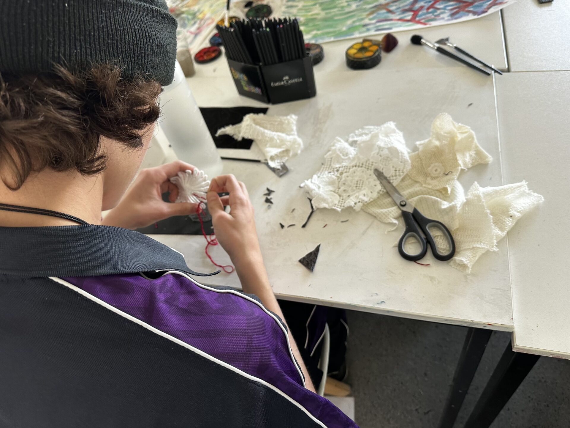 An artist in residence wearing a black beanie and purple collared shirt uses a needle and red thread to stitch pieces of white lace on a table.