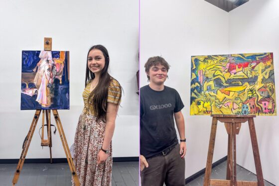A woman stands next to a portrait painting and a man stands by an abstract, brightly coloured work, both displayed on wooden easels in a gallery during an artistic achievement celebration.
