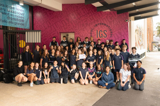A large group of secondary school students posing for a Going the Distance charity fundraiser photo in front of a school building with a pink wall featuring the IGS logo and multilingual text.