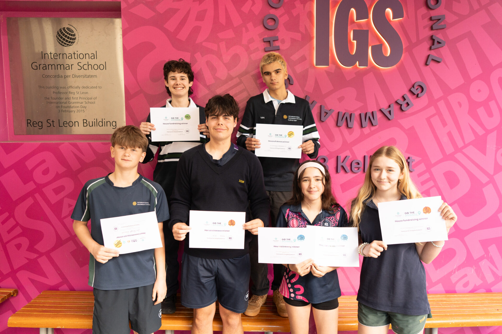Six students in school uniforms stand in a row holding award certificates for a Chris O’Brien Lifehouse charity fundraiser in front of a pink International Grammar School sign.