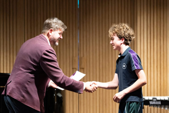 A man in a blazer shakes hands with a student on a concert stage, handing them a certificate.