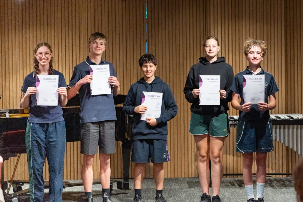 Five students in school uniforms stand in a row holding Certificates of Achievement, looking as prepared for a live music concert as they are for academic success.