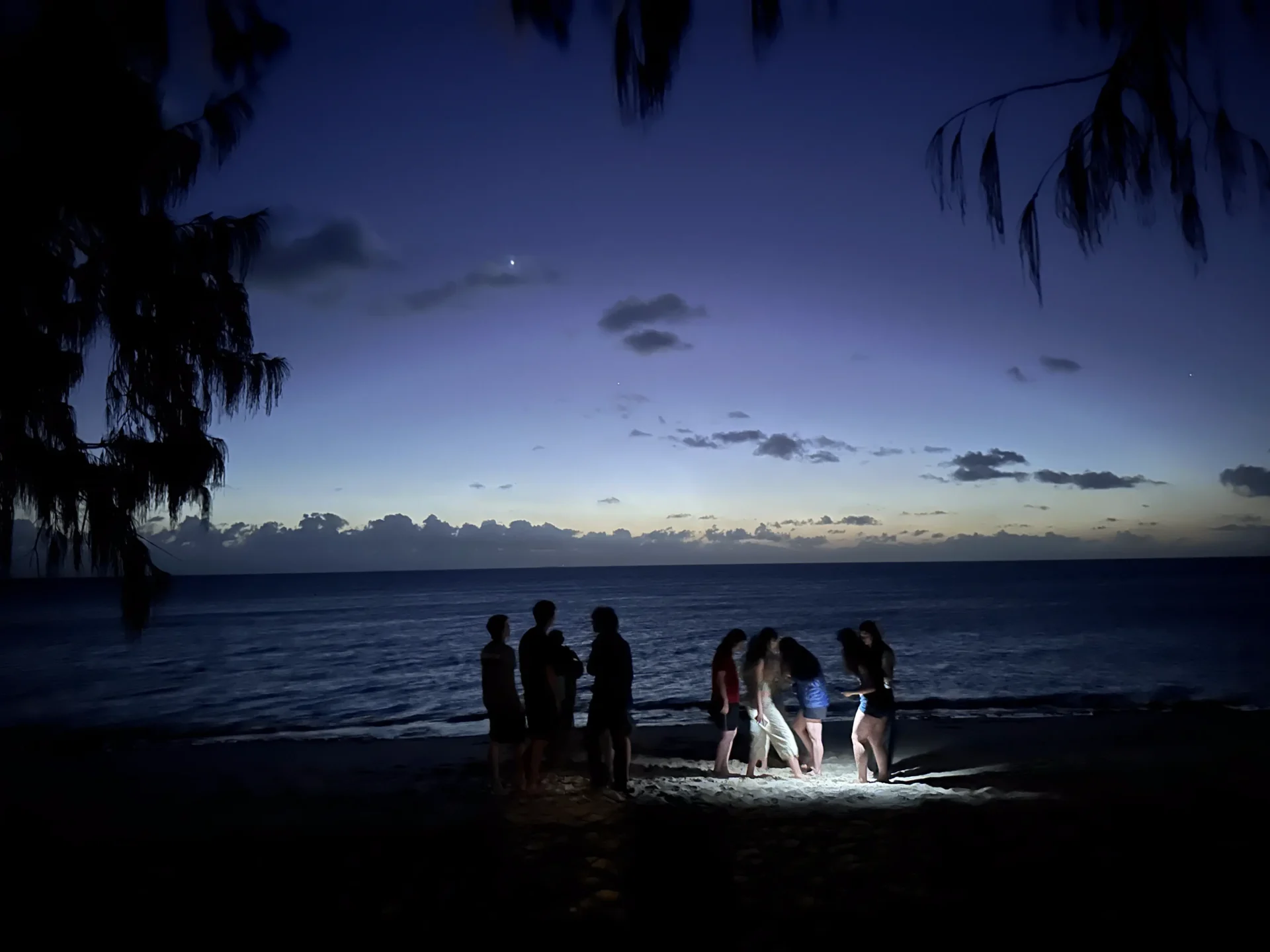 A group of people standing on a beach at twilight, illuminated by a bright light source on the sand.