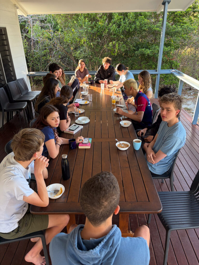 A large group of teenagers sits around a long wooden table on a terrace, eating breakfast.