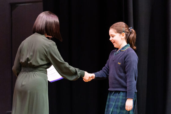 A woman in a dark green dress shakes hands with a schoolgirl holding a certificate on a stage.