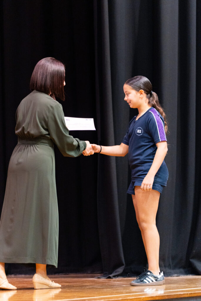 A woman in a green dress shakes hands with a girl in a navy school uniform on stage, celebrating her determination as she hands her a certificate. Black curtains hang in the background.