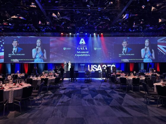 A gala stage with two large screens showing the Finn and Miranda event name and speakers, positioned above a ballroom filled with round dining tables.
