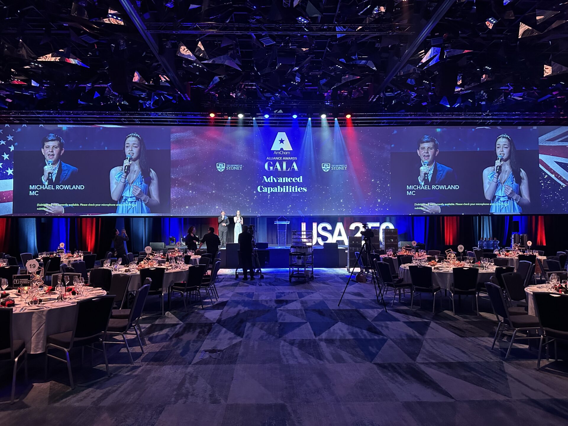 A gala stage with two large screens showing the Finn and Miranda event name and speakers, positioned above a ballroom filled with round dining tables.