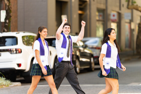 Three students in school uniforms and purple graduation sashes walk across a street during the final school term, with the student in the middle cheering with his arms raised.
