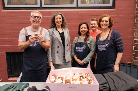 Five people stand behind a table with a box of decorated cupcakes, a sweet gift of thanks; three individuals wear navy blue aprons with the Parents & Carers logo.